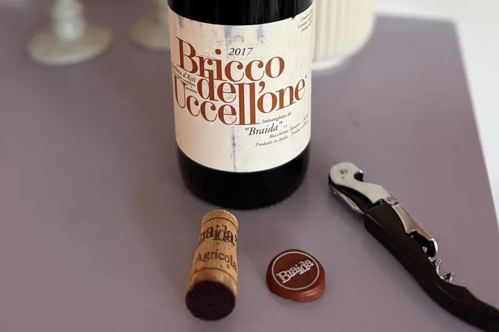 Flasche Braida Bricco dell'Uccellone 2017 mit lesbarem Etikett, daneben Korken, Kapsel und Kellnermesser auf einem lilafarbenen Tisch