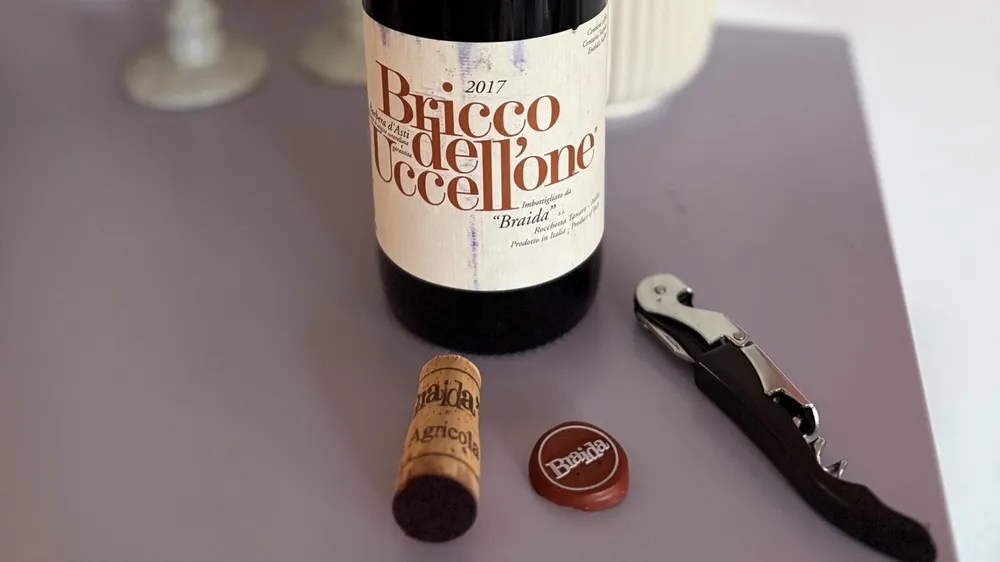 Flasche Braida Bricco dell'Uccellone 2017 mit lesbarem Etikett, daneben Korken, Kapsel und Kellnermesser auf einem lilafarbenen Tisch