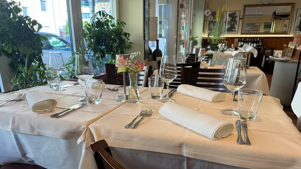 Eingedeckte Tische mit cremeweißen Stofftischdecken, gerollten Servietten und Weingläsern an der Fensterfront des Fellini