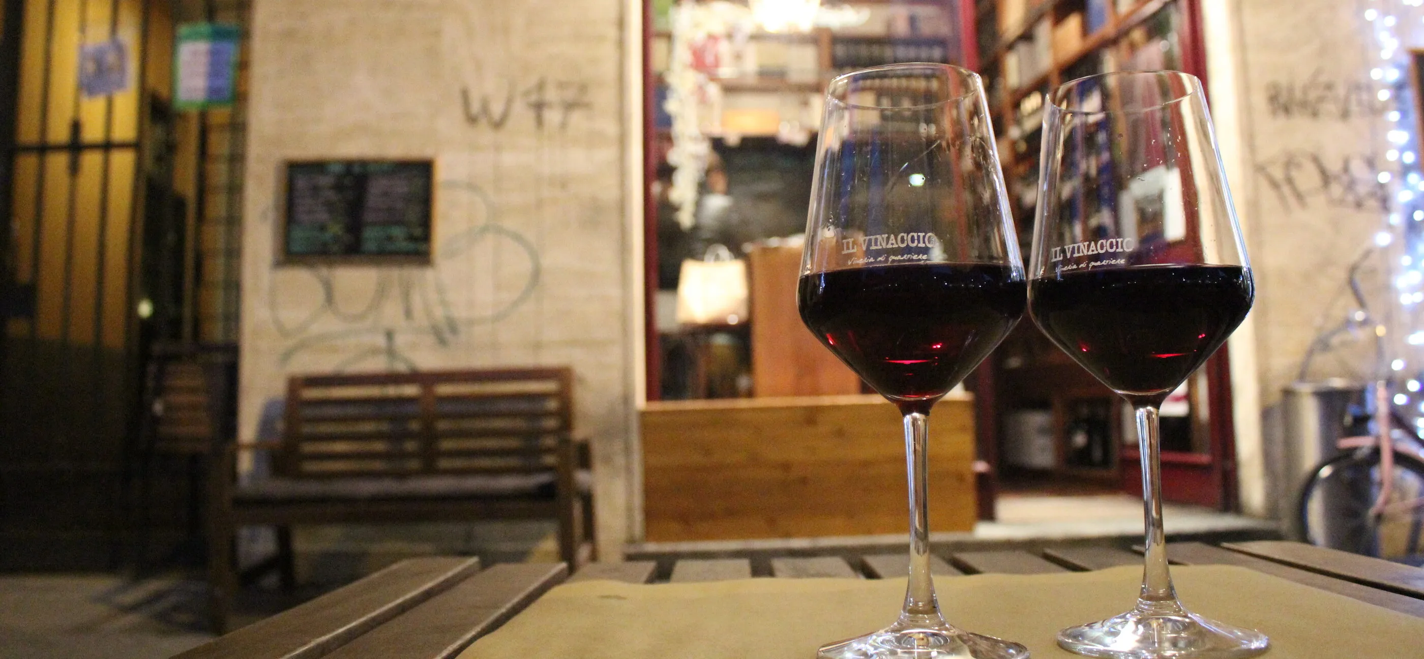 Zwei Gläser Rotwein auf einem Holztisch vor der beleuchteten Fassade einer Weinbar am Abend