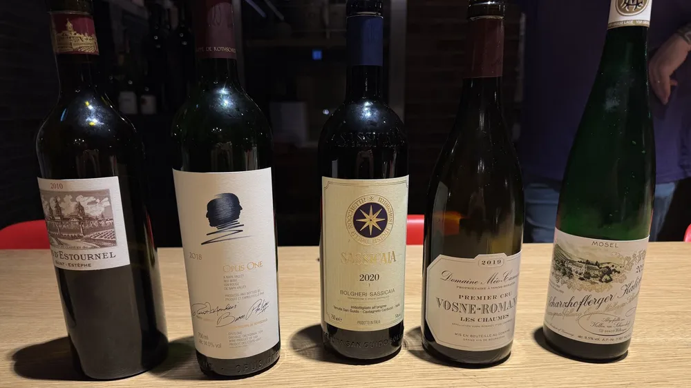 Fünf Weinflaschen nebeneinander auf einem hellen Holztisch: Château Cos d'Estournel 2010, Opus One 2018, Sassicaia 2020, Méo-Camuzet Vosne-Romanée Les Chaumes 2019 und Egon Müller Scharzhofberger Kabinett 2022