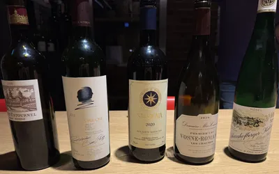 Fünf Weinflaschen nebeneinander auf einem hellen Holztisch: Château Cos d'Estournel 2010, Opus One 2018, Sassicaia 2020, Méo-Camuzet Vosne-Romanée Les Chaumes 2019 und Egon Müller Scharzhofberger Kabinett 2022