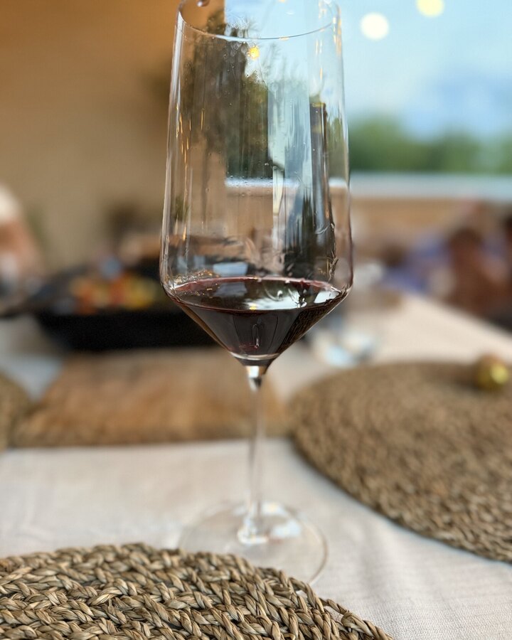 Rotweinglas mit dunklem Wein auf einem gedeckten Balkontisch, im Hintergrund unscharf eine Form mit Essen und eine Lichterkette