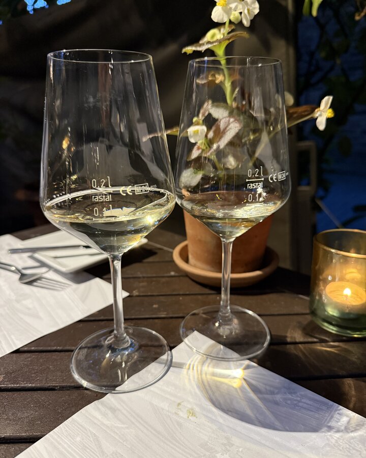 Zwei Weißweingläser mit Eichstrich im abendlichen Innenhof, dahinter Blumentopf und Windlicht