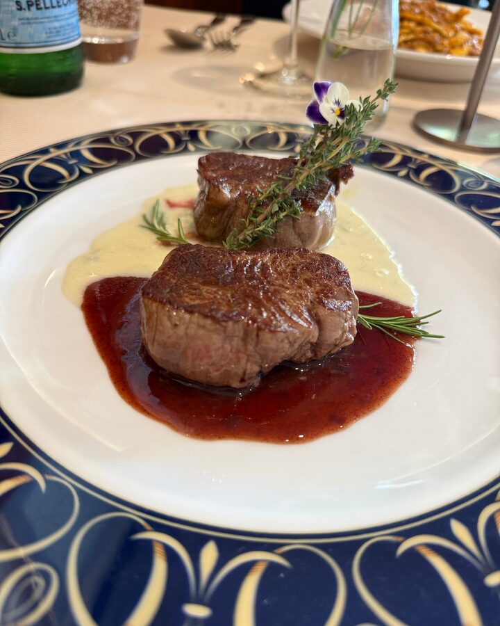 Zwei Filetmedaillons auf dunkler und heller Sauce mit Rosmarin, Thymian und Essblüte auf weißem Teller mit blau-goldenem Rand