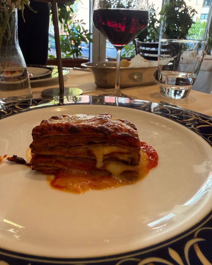 Geschichtete Parmigiana di melanzane mit geschmolzenem Käse und Tomatensauce auf weißem Teller mit blau-goldenem Rand, dahinter ein Glas Rotwein