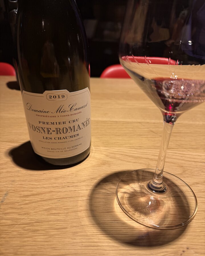 Flasche Domaine Méo-Camuzet Vosne-Romanée Premier Cru Les Chaumes 2019 neben einem Glas Rotwein auf hellem Holztisch
