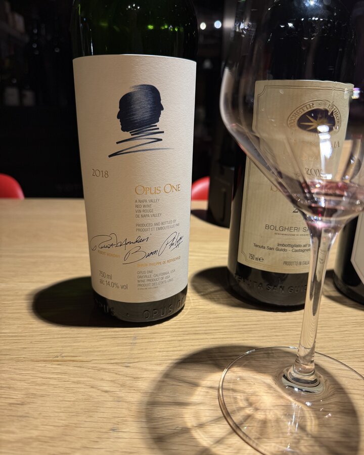 Flasche Opus One 2018 im Vordergrund, dahinter Sassicaia 2020 und ein Weinglas auf hellem Holztisch